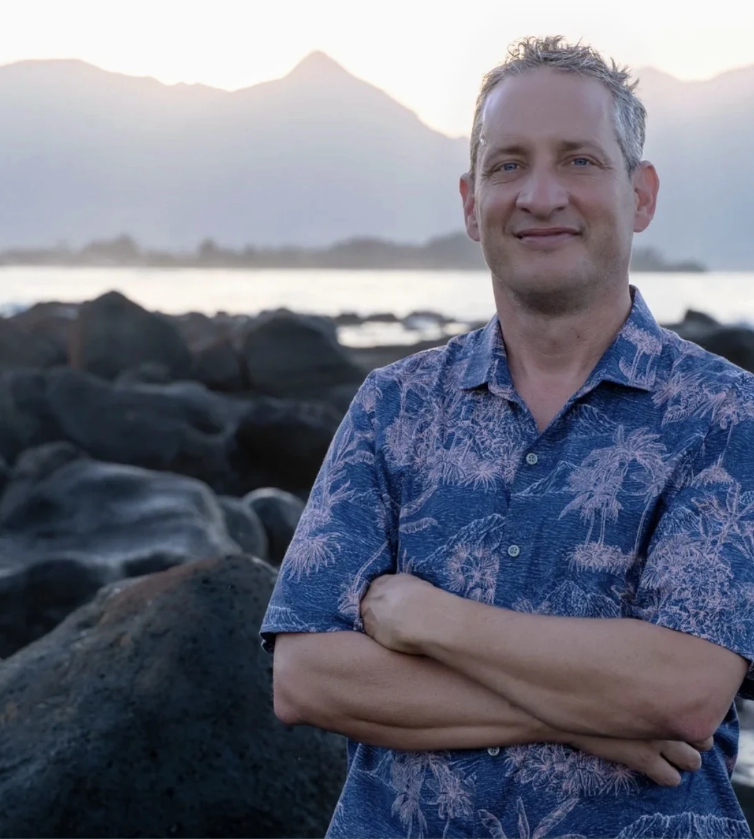 Peter Nathaniel Martin in a blue aloha shirt beside a rocky shoreline.